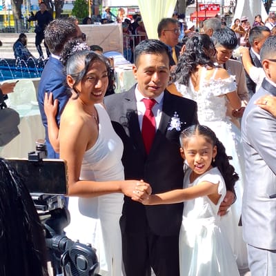 A family is photographed at a wedding in Cerro Colorado, Peru. The bride wears a white dress, and the groom wears a black suit with a red tie. A young girl in a white dress is beside the groom. Other people are in the background, with vendors and tents visible. A smartphone and a camera are in the foreground, capturing the event.