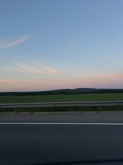 The image shows a landscape scene from the perspective of a moving vehicle on a highway. The sky is a gradient of soft blues and pinks, indicative of either sunrise or sunset, with faint wispy clouds. Below the sky is a rolling landscape of green fields and a dark line of trees forming a distant horizon. A metal guardrail is visible in the foreground, running horizontally across the image, separating the highway from the fields. The highway itself is dark asphalt with a white line marking the lane. The impression is one of traveling through the countryside at dusk or dawn. The location is identified as Kayanovo, Russia.