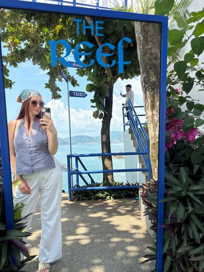 A selfie is taken in a large mirror with a blue frame, showing "The Reef" sign in blue. The woman in the selfie is wearing a gray button-down shirt, white pants, sunglasses, and a patterned headscarf. She is holding a smartphone in her right hand. The mirror reflects a scene with a man on blue stairs, a tree, and a view of a body of water, with foliage visible around the mirror's edge. A directional sign indicates "Hotel" and "Beach." The image suggests a location in Kamala, Thailand, given the coastal setting and tropical foliage.
