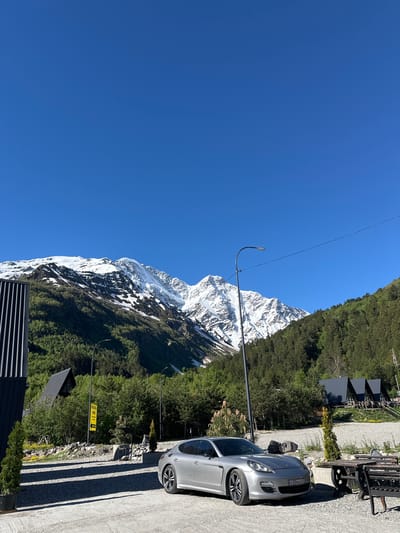 A silver Porsche Panamera is parked on a gravel lot in front of a scenic mountain range with snow-capped peaks and lush green forests. Several modern, dark-roofed buildings, possibly cabins or chalets, are nestled among the trees on the hillside. A street lamp stands near the car, and a picnic table with benches is visible to the right. A yellow sign with Russian text is also present. The sky is clear and blue. The location is the city of Terskol, Russia.