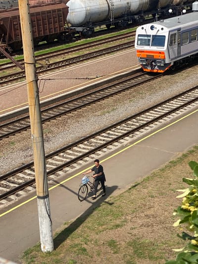 In Romodan, Ukraine, a person is walking a bicycle on a paved platform next to train tracks. The person is wearing a black shirt and black pants, and they are pushing the bicycle which has a blue and white bag in the basket. The bicycle is leaning slightly to the right. Train tracks run parallel to the platform, and there are several freight cars and a passenger train visible in the background. The ground is covered with gravel and grass. A concrete pole with electrical wires runs vertically along the left side of the image. The scene appears to be outdoors on a sunny day.