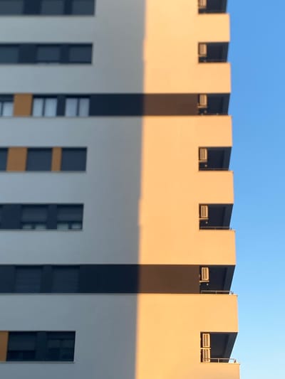 The image displays a close-up, abstract view of a modern apartment building, likely situated in the Cukarica Urban Municipality, Serbia, given the context. The building exhibits a minimalist design with white and dark grey horizontal bands punctuated by rectangular windows. One side of the building is illuminated by the warm glow of sunlight, creating a stark diagonal shadow that divides the facade. On the sunlit side, balconies are visible, each equipped with visible air conditioning units. The sky above is a clear, vibrant blue. The composition focuses on the architectural elements and the play of light and shadow, creating a geometric and serene urban landscape. There are no people or animals visible in the image.