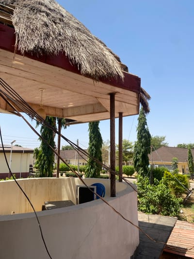 The image captures an exterior scene under a clear blue sky in Tudun Nupawa, Nigeria, viewed from an elevated vantage point. In the immediate foreground, a curved, light-colored concrete parapet wall with visible surface cracks partially encloses an area. Within this enclosure, a dark-colored rectangular object and a blue plastic chair are discernible. Multiple black cables descend from the upper left, crossing the frame diagonally and resting on the parapet wall.
Above the parapet, a structure features a prominent thatched roof composed of dry straw, supported by a dark red fascia board and a light-colored, paneled underside. Two textured brown columns support this roof structure. A single, light-colored pendant fixture hangs from the paneled underside.
Beyond the immediate structure, the environment includes multiple tall, slender green trees and various green foliage. In the middle distance, several single-story buildings with light-colored walls and brown-tiled roofs are visible, interspersed among the greenery. A solar-powered street light and utility poles with overhead wires are present on the left side of the background. No individuals are visible within the frame.