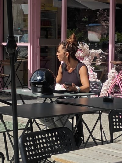 A woman with her hair in a bun sits at an outdoor table in front of a pink building. She is wearing a dark sleeveless top and is holding a smartphone in her right hand. A black motorcycle helmet is on the table in front of her. Behind her, a pink bicycle is parked next to a decorative display of white flowers. The building appears to be a cafe or shop with large windows. The scene is set in Molenbeek-Saint-Jean - Sint-Jans-Molenbeek, Belgium.
