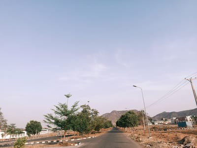 The image depicts a road in Rafin Pa, Nigeria. The road is paved and stretches into the distance, lined with trees on either side. A mountain range is visible in the background under a clear sky. Buildings are seen in the background. Utility poles and street lights are present along the side of the road. The area around the road appears to be undeveloped or under construction, with dirt and sparse vegetation. There are no people visible in the image.