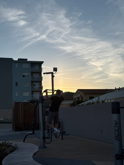 A man is performing pull-ups on an outdoor gym apparatus at sunset in Vallecrosia al Mare, Italy. The man, silhouetted against the sky, wears a dark t-shirt and shorts. The gym equipment is positioned in front of a tall, light-blue apartment building. In the background, other buildings with tiled roofs are visible, and the sky is filled with wispy clouds and contrails, indicating the setting sun. The ground is paved with gravel and features a curved wooden seating area. A small airplane is visible in the distance.