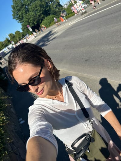 The image shows a woman taking a selfie outdoors on a sunny day. She is wearing sunglasses, a white shirt, and a small green bag. In the background, there are people walking and cycling, a road, trees, and buildings. There is also a shadow of the woman on the ground. Based on the presence of what appears to be a temporary barrier with traffic cones and a sign that resembles a "no parking" sign with German lettering, it is possible the scene is related to an event or construction near the Kiel Fjord, possibly along the Düsternbrooker Weg or a similar waterfront promenade in Kiel, Germany. The overall impression is a casual outing in a public urban space.