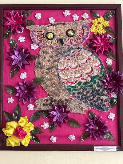 The image displays a framed handicraft, a decorative artwork featuring an owl, constructed from various textured materials such as jute, yarn, and fabric ribbons. The owl is depicted with large, button-like eyes and a textured body, with its wings adorned with intricate patterns of ribbons and yarn in shades of brown, beige, blue, and green. The background of the artwork is a vibrant pink fabric. Surrounding the owl are numerous fabric flowers and leaves, made from satin ribbons in shades of pink, purple, and yellow, as well as some small white flowers and dark green leaves. At the bottom of the frame, there is a white label with text in Cyrillic script, which appears to be a title or description of the artwork. The artwork is displayed on a light-colored wall. Based on the provided label information, the artwork is titled "Magic Flowers" and is associated with a territorial social service center for the population of the Polotsk district, likely a craft created by individuals from the Vetrynsky rural council in Belarus.