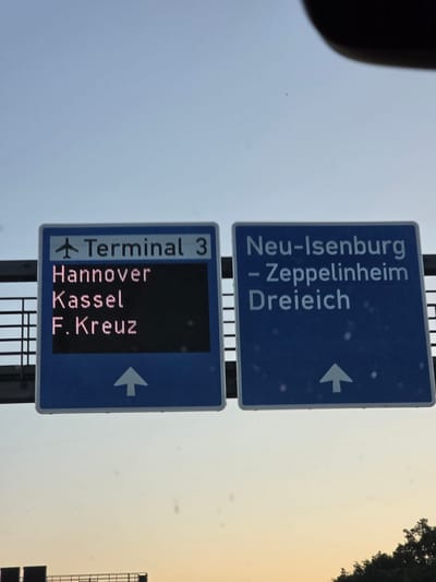 The image shows two blue directional road signs against a pale sky. The sign on the left indicates "Terminal 3" with an airplane icon, followed by a list of destinations: "Hannover", "Kassel", and "F. Kreuz", with an upward arrow below. This sign appears to be a digital display with illuminated pink text. The sign on the right lists destinations "Neu-Isenburg", "- Zeppelinheim", and "Dreiich", also with an upward arrow below. Both signs are mounted above a grey metal structure, possibly a highway barrier or bridge. The sky suggests it is either early morning or late evening. The sign for Zeppelinheim places it within the context of directing traffic towards this location.