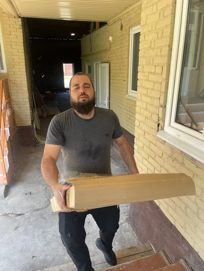 A bearded man with closed eyes, appearing fatigued or stressed, carries a large cardboard box. He is wearing a dark grey t-shirt with visible sweat stains and black athletic pants. The man is walking up a set of wooden steps, suggesting he is entering or leaving a building. The surroundings include a weathered brick building with light yellow paint, a covered walkway with a corrugated metal roof, and a darker, open structure in the background that appears to be a garage or shed with a doorway leading outside. The ground is concrete with some cracks. The scene is set outdoors, likely in a residential or industrial area. The presence of wooden planks and construction materials in the background hints at ongoing work or storage.
