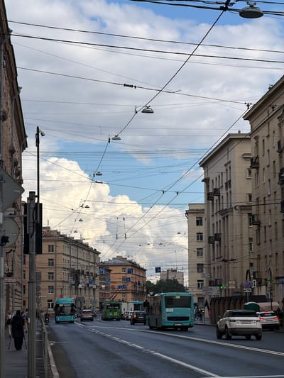 The image depicts a street scene in what appears to be a city, with a focus on overhead wires and buildings. On the right side of the street, there are several multi-story buildings with a light-colored facade. These buildings have numerous windows and balconies. The street itself is wet, suggesting recent rain or cleaning. Several vehicles are visible on the road, including buses and cars. A light-colored SUV is prominently parked on the right side. Overhead, a complex network of wires crisscrosses the sky, likely for public transportation like trolleys or trams, and also for electricity. The sky is partly cloudy with large white clouds. On the left side of the street, there are more buildings, a pedestrian sidewalk with a few individuals, and a tall lamppost with traffic signals and cameras. The overall impression is a busy urban environment. The location of "Dnp Ryzhiki, Russia" is not explicitly identifiable from the visual cues in the image.