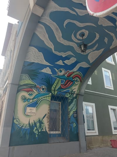 The image shows a street view in Aveiro, Portugal, with a building facade decorated with a large mural. The mural features two stylized birds with prominent eyes and red and blue markings, surrounded by green foliage. Above the birds, the mural depicts a blue sky with swirling white clouds and a vintage-style street lamp. A sign reading "Arco do Comércio" is integrated into the mural design. Above the mural, there are windows of a green building. A road sign is also visible.