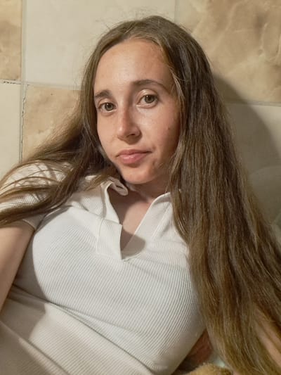 The media file is a close-up, portrait-style photograph of a young woman with long, brown hair. She is lying down, with her head tilted upwards and her body angled such that her face is the primary focus. She is wearing a white, ribbed top with a collar. Her hair falls loosely around her shoulders and across her chest, obscuring some of her upper body.

Her facial features are visible: she has light brown eyes, a small nose, and full lips that are slightly parted. Her skin appears fair with some freckles and slight redness. The lighting suggests it is daytime, possibly indoors due to the tiled background, which appears to be a light beige or cream color with some darker, marble-like veins. The tiles are arranged in a grid pattern and show some signs of wear and tear, with visible grout lines and a slight crack in one of the tiles.

The woman's expression is neutral, with her gaze directed slightly off-camera. Her hair is a dominant element in the composition, framing her face and extending beyond the edges of the frame. The overall impression is casual and intimate, likely a selfie. There is no visible text in the image. The location context provided is Vypasne, Ukraine.