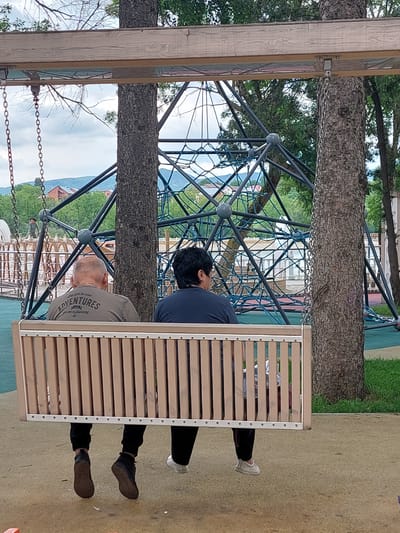 In an outdoor playground setting, two individuals are seated on a wooden swing. The scene appears to be in the vicinity of Аликоновка, Russia, judging by the surrounding vegetation and architectural styles. One person, facing away from the camera, has short gray hair and is wearing a gray t-shirt with visible text on the back. The text appears to say "ADVENTURES" in large letters, with smaller text below that reads "go on a journey". This person's legs are visible, wearing dark pants and brown shoes.

The second individual, also facing away, has dark, short hair and is wearing a dark blue t-shirt and black pants. Their legs are also visible, wearing white shoes. They are seated to the left of the first person on the swing.

The swing itself is constructed with a wooden frame and a slatted wooden seat. It is suspended by chains from a larger structure, possibly part of a climbing frame. Behind the swing, a complex network of metal bars and blue netting is visible, suggesting a climbing apparatus. Trees with rough bark surround the area, and in the background, hints of buildings and hilly terrain can be seen under a bright sky, suggesting it is daytime with fair weather. The overall mood seems relaxed, with the individuals enjoying a moment of rest on the swing.