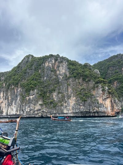 An image shows a wide, slightly low-angle view of a large, rocky island with green vegetation covering its top. The island has sheer, gray-brown cliffs with vertical striations, and some patches of orange and red rock. The water in the foreground is dark blue and choppy with white foam. Two longtail boats, common in Thailand, are visible on the water. One boat, closer to the viewer and on the left, has a visible engine and appears to be moving. Another longtail boat with a blue canopy and passengers is further away, to the right of the island. The sky is overcast with gray clouds. The scene is likely a coastal area in Thailand, possibly near Phi Phi Islands, given the characteristic limestone cliffs and longtail boats.
