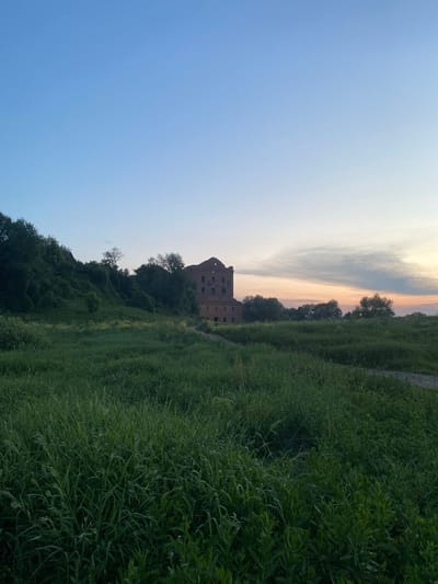 The image captures a serene landscape in Yuryatino, Russia, with an old brick building at its center. The sky is a gradient of light blue fading into a soft pink and orange hue near the horizon, indicative of dusk. In the foreground, tall green grass stretches across the frame, creating a lush, natural carpet. To the left, a sloping hill covered in dense green foliage rises, contrasting with the open field. The brick building, appearing somewhat dilapidated with visible damage and missing sections, stands on the right side of the hill, its architecture suggesting it might be an old mill or industrial structure. The overall atmosphere is peaceful and slightly melancholic, evoking a sense of history and natural beauty. There are no people or animals visible in the image.