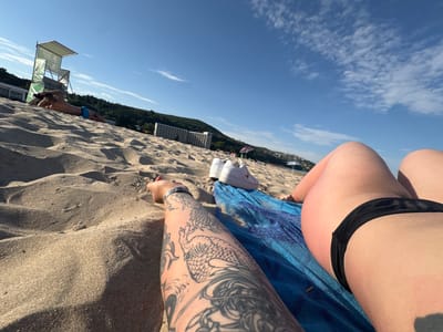 This is a first-person perspective photo taken on a sandy beach. The foreground shows a person's tattooed arm and leg, as well as a blue towel and white sneakers. In the mid-ground, another person is lying on the sand near a lifeguard tower. In the background, a large hotel building and a hillside covered in trees are visible. The sky is blue with some scattered clouds, indicating it is daytime. The overall scene suggests a relaxed day at the beach. There is no visible text in the image.