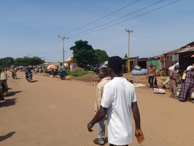 In Kakuri Gwari, Nigeria, a bustling marketplace scene unfolds on a dusty, unpaved road under a clear blue sky. The road is lined with makeshift stalls constructed from corrugated metal and wood, displaying various goods, including sacks of produce and baskets. Several individuals are present, some walking along the road, others engaged in commerce. A few motorbikes are visible, with one person riding and another parked on the side. Overhead, power lines crisscross the sky, supported by utility poles. The overall environment suggests a lively and active market day, characterized by its informal setup and the presence of everyday commerce.