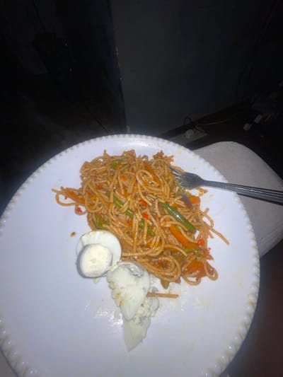 A plate of spaghetti with vegetables and sliced boiled eggs is on a white decorative plate. A silver fork is partially visible, resting on the spaghetti. The scene is dimly lit and the surrounding area appears to be dark, though a portion of a chair or couch is visible behind the plate. There is no indication of location in Kurunduma, Nigeria or any other identifying information in the image.