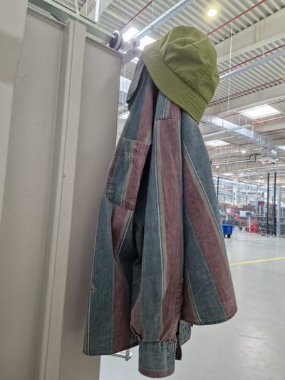 A green bucket hat is perched on top of a folded striped shirt. The shirt has alternating stripes of dark blue and a reddish-brown color, with thin white stripes interspersed. The shirt appears to be made of a denim-like material. The clothing is resting on a white railing or shelf, which is part of a larger structure.
In the background, a large industrial or warehouse-like space is visible. The ceiling is high and features exposed metal beams and red pipes. Skylights allow natural light to filter into the space. Rows of shelving units are visible in the distance, some holding boxes and other items. Forklifts or similar industrial vehicles can also be seen, suggesting an active work environment. The floor is a light gray concrete. A yellow line is painted on the floor, possibly indicating a walkway or safety zone. The overall setting suggests a logistics center, factory, or distribution warehouse.
The image is taken from a slightly low angle, looking upwards towards the clothing and then into the background. The lighting appears to be a mix of natural light from the skylights and artificial lighting within the building. There is no indication of the time of day beyond the presence of daylight. No people are clearly visible. The scene appears to be utilitarian and functional.
