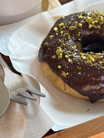 The image displays a close-up, elevated view of a single chocolate-frosted donut topped with crushed pistachios, resting on white parchment paper. The donut, positioned prominently in the right half of the frame, is dark brown with a glossy, irregular chocolate coating and numerous light green pistachio pieces scattered across its surface. Visible along its bottom edge is the lighter, beige-colored donut base. To the left, a silver-colored metal fork with three tines and a knife with a serrated edge are partially visible, lying on the parchment paper. The paper exhibits minor creases and a faint reddish smudge near the cutlery. These items are situated on a light brown wooden surface. A portion of a white ceramic cup or bowl is visible out of focus in the upper left corner. The scene is set in San Phra Net, Thailand, and suggests a prepared dessert or snack. No individuals are visible.
