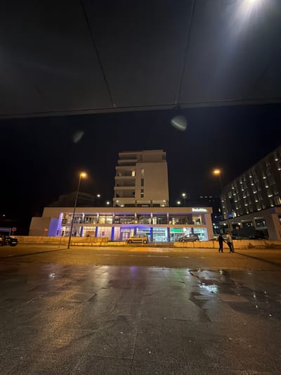 At night, a partially lit street showcases buildings. "Deloitte" is visible atop one building, next to a medical office. A modern, multi-story structure with open spaces stands nearby. Streetlights illuminate the wet pavement, creating reflections. Two figures stand on the sidewalk. A tall building is partially visible in the background, while a dark structure looms on the left.