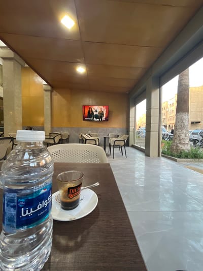 This media file captures an indoor cafe or restaurant setting, looking out onto a street scene through large windows. In the immediate foreground, on a dark brown wooden table, sits a clear plastic water bottle with a white cap and a glass containing remnants of coffee, placed on a white saucer with a spoon.

Beyond the table, the cafe interior features light brown wood-paneled walls, with two square, glowing light fixtures mounted high up. A flat-screen television is affixed to one wall, displaying a scene with two individuals and the English text "REGRETTING YOU." Several white and black chairs are visible, arranged in what appears to be seating areas. The floor is covered in light grey or off-white tiles, some showing reflections.

The large windows offer a clear view of the outside. A prominent palm tree with a thick trunk stands on the street. Behind it, light-colored buildings line the street, where cars and motorcycles are parked, and a person can be observed. The bright natural light streaming in suggests it is daytime.

Visible text includes Arabic script on the water bottle: "كوافينا" (Quafina), "ماء المائدة" (Table water), and "قليل الصوديوم" (Low sodium), along with numbers "33" and "001". The coffee glass features a "DICAR" logo and the text "To taste," while the saucer has "CAFE" printed on it. The scene's details, particularly the Arabic text and the palm tree, are consistent with the location context of Sebt Gzoula, Morocco.
