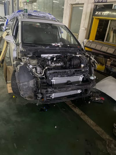 The image shows a white car with its hood open, parked inside a garage or repair shop. The car's front bumper and some of its internal components are disassembled. Several parts are lying on the ground around the vehicle. The garage appears to be industrial, with concrete floors and exposed walls. In the background, there are shelves, machinery, and signage, including the "ZETA" logo. The overall scene suggests that the car is undergoing repairs. The location is identified as Panyu District, China.