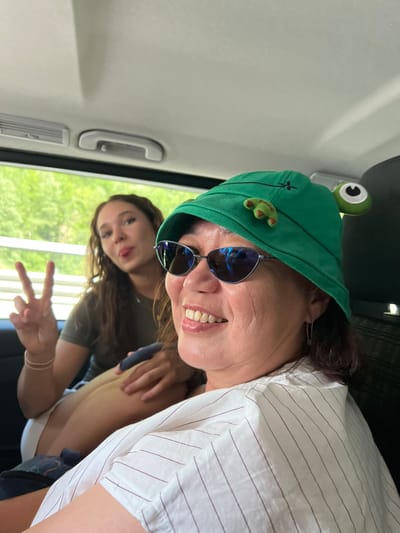 Two individuals are seated inside a vehicle, with the foreground dominated by a woman wearing a green bucket hat adorned with googly eyes and a frog appliqué. She also sports sunglasses and a white pinstriped shirt. Her mouth is open, revealing teeth, and she appears to be smiling. Behind her, a younger woman with long, curly brown hair is visible, sticking out her tongue and making a peace sign with her fingers. She is wearing a dark green t-shirt. The background shows greenery through the car window, suggesting an outdoor setting. The interior of the vehicle is dimly lit, with a light-colored ceiling and a grab handle visible. The scene does not provide any specific visual cues to pinpoint its location within the city of Les Houches, France.