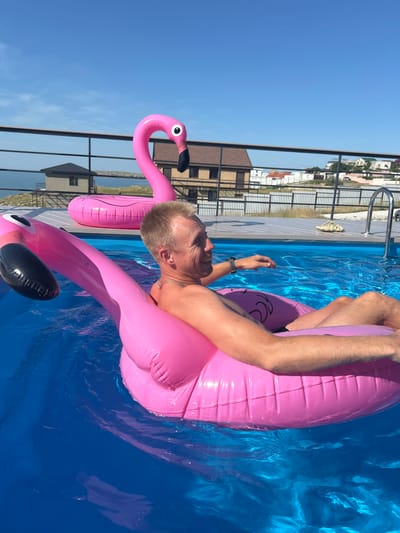 In this medium shot, a man is relaxing in a bright blue swimming pool, floating on a large, inflatable pink flamingo. The man is leaning back on the flamingo, his head resting on its body, and his arms are spread out. He is wearing a silver chain around his neck and a watch on his left wrist. His expression appears content and relaxed.
The flamingo is a prominent object, its neck curved upwards and its body forming a comfortable float. The water is clear and reflects the bright sunlight, creating a shimmering effect. In the background, a fence and some buildings are visible, suggesting an outdoor setting. The sky is a clear, vibrant blue, indicating a sunny day. The overall impression is one of leisure and enjoyment on a warm day. The location context provided suggests this scene is taking place in Mysivske rural settlement, Ukraine.