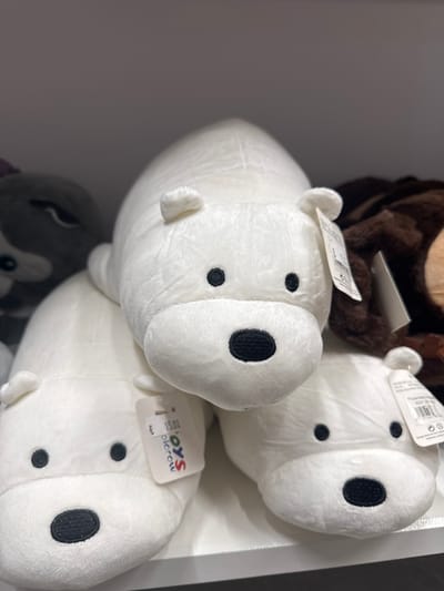 The image displays a close-up of several plush toys, primarily white with black embroidered features. The toys appear to be stylized animals, possibly seals or bears, with round bodies and distinct facial markings. One toy in the foreground is predominantly visible, showing its snout and two black oval eyes. To its right and slightly behind it, another similar white plush toy is partially visible. A dark brown plush toy, possibly a bear, is visible at the top of the frame, behind the white toys. The toys are arranged on a white surface, likely a shelf or display area in a retail setting.

Visible text includes a tag on the foremost white toy that reads "TOYS plorow" with a price tag showing "€ 15.00". Another tag is visible on the toy behind it. The overall scene suggests a retail environment where these plush toys are being sold. The lighting is consistent with indoor artificial lighting. The context provided indicates the location is Keila, Estonia, suggesting these are items available for purchase in that region. No people are visible in the image.