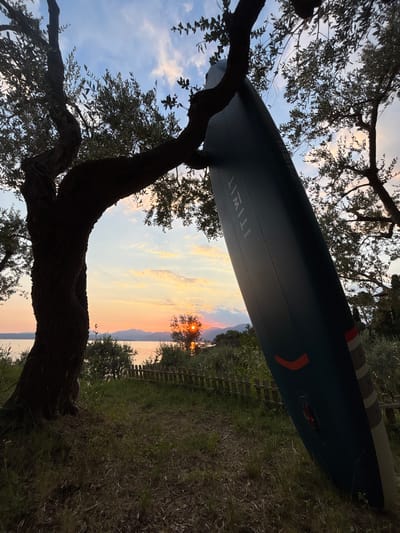 A dark, textured paddleboard leans against a twisted, silhouetted olive tree. The paddleboard is a deep teal color with a raised, textured surface. The name "ITIWIT" is visible in white lettering near the top of the board. The tree's branches reach out, framing a sunset over a body of water. The sky is a gradient of soft orange, pink, and blue, with scattered clouds. In the distance, silhouetted mountains form a distant horizon line. The foreground consists of grassy terrain and dark soil. A rustic wooden fence runs along the edge of the water. The scene appears to be a serene outdoor setting, likely near Lake Garda, Italy, given the mention of the city. No people are visible in the image.