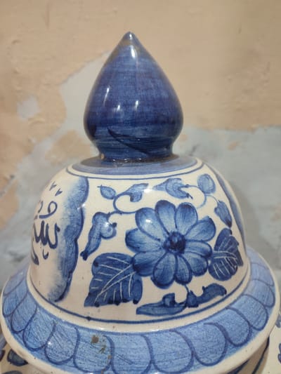 The image displays a close-up, eye-level view of the top portion of a white ceramic vase with blue floral and Arabic calligraphy designs. The vase has a conical blue lid. The primary subject is the decorative ceramic vase, with its intricate blue painting. The background consists of a textured, light-colored wall, possibly plaster, with areas of peeling paint. The wall's texture and the overall lighting suggest an indoor setting, potentially in a home or a storage area. There is no activity or event depicted, as the image focuses solely on the object.
Notable details include the blue and white color scheme, characteristic of traditional ceramics. The blue paint on the vase features floral motifs, including a prominent, large flower with detailed petals, and smaller leaves and vines. To the left of the flower, there are strokes of blue paint that resemble Arabic calligraphy. The ceramic itself shows signs of crazing, a network of fine cracks on the surface of the glaze, which adds to its aged or artisanal appearance. The conical lid is a solid, glossy blue. The background wall is uneven and textured, with patches of lighter and darker tones, indicating wear or an unfinished surface. The lighting appears to be ambient, with soft shadows and highlights on the vase, indicating it's not direct sunlight. No specific time of day or weather conditions can be inferred. The location context provided is Blang Cut, Indonesia, but no specific visual cues within the image confirm this location. There are no people or animals present. The image is static, focusing on the object itself and its decorative elements.
