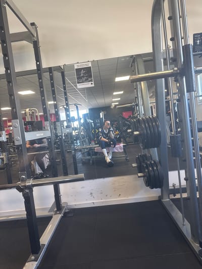 The image shows the interior of a gym in Neuville-aux-Bois, France. The space contains various weightlifting machines and equipment. A person is visible seated on a weightlifting machine, facing away from the camera, with a cell phone in hand. The individual appears to be taking a picture. Multiple weight plates are loaded onto a machine. The gym has a tiled floor and fluorescent lighting. A poster with instructions or information in French hangs on the wall. Other gym equipment is seen in the background.