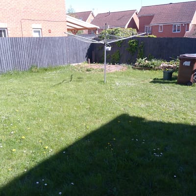 This image captures a sunny residential backyard or garden in Corby, United Kingdom. The foreground features a lush green lawn, liberally sprinkled with small white daisies and bright yellow dandelions. A prominent, dark triangular shadow, cast by an unseen object, extends across the bottom right section of the lawn, indicating bright sunlight.
In the midground, a silver rotary clothesline stands in the center of the lawn, equipped with multiple drying lines. A small white bucket is clipped to its central pole. Behind the clothesline, a dark grey or black wooden slatted fence serves as a boundary, with some sections partially covered by climbing green foliage. To the right, there's an unkempt area that appears to be a raised garden bed or patch of disturbed earth with some plants and debris. A large brown wheelie bin with wheels is positioned to the right, bearing an unreadable light-colored sticker or label. A smaller green plastic bin or bucket sits on the grass near the wheelie bin.
The background reveals several brick houses with distinctive reddish-brown tiled roofs and white-framed windows, characteristic of UK residential buildings. On the far left, a wooden pergola-like structure with a translucent roof extends from one of the houses. The sky is clear and bright, indicating good weather. No people are visible, and the scene depicts a quiet, everyday garden on a sunny day.