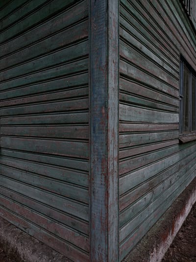 The image shows the weathered exterior of a wooden building, likely a shed or an old house, in the Ракітніцкі сельскі Савет, Belarus. The building's walls are constructed from horizontal wooden planks painted a dark, desaturated green, which is peeling in many areas, revealing reddish-brown underlayers. A vertical wooden beam, also showing signs of wear and peeling paint, forms a corner of the structure. To the right, a small window with a dark frame is visible, set into the wooden siding. The base of the building rests on a rough, concrete-like foundation, which meets the ground covered in dry leaves and debris. The overall impression is one of age, neglect, and the passage of time, with the muted colors and visible decay suggesting an outdoor setting in a rural or semi-rural environment. There are no people or animals present in the image.