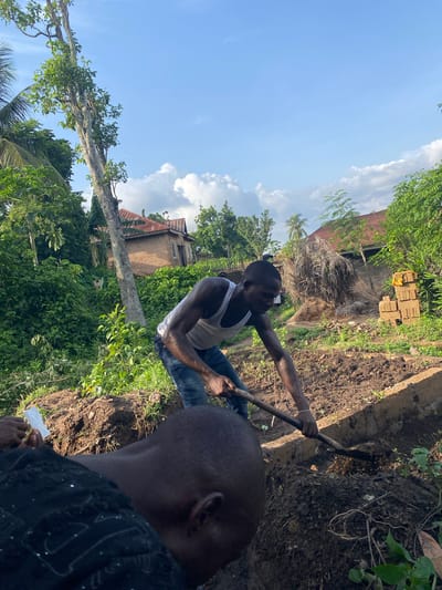 An eye-level, medium shot captures a man in a white tank top and blue jeans digging into the earth. He is using a tool with a long handle, possibly a shovel or hoe, to excavate the soil. The surrounding environment indicates an outdoor setting with lush greenery, including trees and vegetation, as well as a building with a corrugated red roof visible in the background. In the foreground, another man partially obstructs the camera's view. The scene is lit by natural daylight, and the sky is visible at the top of the frame. The image was taken in the city of Unwana, Nigeria.