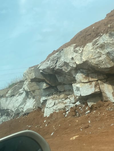 The image displays a rugged rocky cliff face, viewed from a perspective that suggests it was taken from a vehicle. The cliff, composed of large, fractured grey and off-white rocks, dominates the upper part of the scene. Above the main rock formation, a distinct layer of reddish-brown earth or soil is visible, scattered with small white fragments. Below the cliff, a wide expanse of bright, clear blue sky indicates daytime conditions.
In the bottom right corner, a partial view of a curved, grey metallic object, likely part of a vehicle's interior like a dashboard or rearview mirror housing, is visible. This detail suggests the photograph was captured while moving through the landscape of Gosa, Nigeria. Thin, dark lines, possibly power lines or similar cables, can be seen against the sky, stretching horizontally across the lower-middle section of the image.
Notable details on the rock face include numerous cracks and fissures, highlighting its weathered texture. Near the center of the rock formation, on a lighter-colored section, black markings are present. These appear to be graffiti or symbols; one distinct mark resembles a stylized number "2" or "Z," possibly followed by other indistinct characters, or an "S" with an arrow. The scene captures a natural geological feature alongside human elements, providing a glimpse of the environment in Gosa.