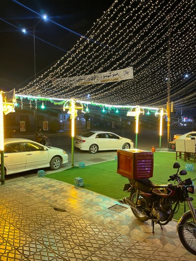 The image depicts a nighttime scene in Kabul, Afghanistan, likely a restaurant or a similar establishment, adorned with numerous string lights. The lights are strung across overhead, creating a canopy effect, and also illuminate poles with green and yellow decorative elements.

In the foreground, a tiled walkway leads into the scene. To the right, a motorcycle with a red delivery box on the back is parked. Beyond the walkway, an area covered in artificial turf is visible, with two white cars parked on it. One car is a sedan, and the other is partially visible. There are also stacks of water bottles on the turf.

In the background, buildings and streetlights are visible. A banner hangs across the overhead lights with Arabic script. A person can be seen on a bicycle in the middle ground, riding on what appears to be a paved road behind the parked cars. The overall atmosphere is that of an outdoor dining or gathering space illuminated for evening use.