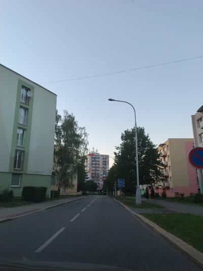 The image depicts a street scene in Prachatice, Czechia. On the left, a pale green apartment building with multiple windows and balconies runs parallel to the street. To the right of this building, a tall, mature deciduous tree stands, partially obscuring a yellow apartment building. Further to the right, a taller apartment building with a grey and red facade is visible, with a pink and beige building in front of it. A traffic light with a red circle and a blue diagonal bar is mounted on a pole on the right side of the road. Two individuals are walking on the sidewalk near the pink and beige building. The street itself is paved and has white lane markings. The sky is clear and light blue, suggesting it is either early morning or late afternoon.