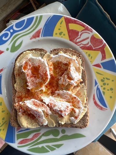 A single slice of whole wheat bread, generously spread with hummus, topped with dollops of what appears to be ricotta cheese, and sprinkled with paprika, sits on a white plate with a multicolored geometric pattern. The plate rests on a surface with potential sunlight. The location is Urbanización Encinares De Alcolea, Spain. No people are visible.