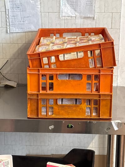 The image depicts a retail or backroom area within a commercial establishment in Ihtiman, Bulgaria. The central focus is a stack of three orange plastic crates, branded "Кума" (Kuma), resting on a brushed metal countertop. The top crate is tilted, and its front edge displays molded text in Cyrillic, "ТАБЛИЦА ЛОТОРИЯ" (Lottery Board). This crate is filled with numerous bundles of lottery scratch cards or tickets, many prominently featuring the word "WIN" in English. The underlying two crates also contain various packaged items, visible through their open rectangular sections, consistent with more lottery products or small retail goods.

Behind the crates is a wall clad in small, rectangular white tiles. Affixed to this tiled wall are two pieces of white paper with handwritten notes in Cyrillic (Bulgarian), appearing to be lists or schedules. The metal countertop extends across the lower half of the image, displaying a black Monster Energy logo sticker and several other smaller, indistinct stickers. To the left on the countertop, a coiled black cable and a folded white cloth are visible. At the bottom left, a portion of a black object, likely an armrest of a chair or stool, is visible. Adjacent to it, a white plastic container is partially visible, bearing handwritten Cyrillic text, including "САМО СЕ ПОДДЪРЖАЙ" (Just maintain yourself) and "ББЛ 2,000". No people are present in the scene, and there are no discernible actions or interactions beyond the static arrangement of objects. The lighting suggests an indoor, artificial light source.