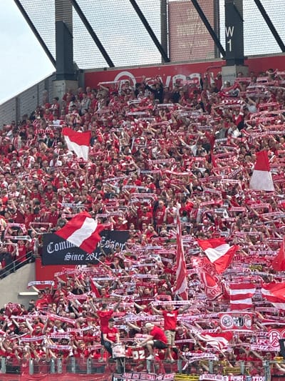 This image captures a dense crowd of soccer supporters, likely at a stadium in Essen, Germany, judging by the German-language banners and flags. The fans are predominantly dressed in red and white, and many are waving scarves and flags of the same colors. A large banner with the words "Comm since 2012" and "Essen" is visible. The crowd appears to be engaged in a passionate display of support, cheering and waving their gear. A portion of the stadium structure, including a metal framework and a sign with "W1" visible on it, is seen in the background.