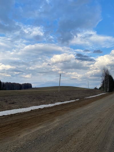 The image depicts a vast, open rural landscape in Nekrasovo, Russia, characterized by a gravel road, fields, distant trees, and a dynamic sky. The scene appears to capture the transition from late winter to early spring.
In the foreground, a wide, unpaved gravel road extends diagonally across the frame, from the lower right towards the middle left. A narrow, elongated strip of melting snow runs along the left edge of this road, indicating recent cold weather or remaining snow patches. Adjacent to the road and snow is a large, seemingly barren field with light brown, dry vegetation, stretching towards the horizon.
Further into the mid-ground, two wooden utility poles with power lines are visible, providing a human-made element within the natural setting. Beyond the field and poles, a dense line of trees forms the distant horizon. These trees appear to be a mix of bare deciduous trees and dark evergreen conifers.
The sky dominates the upper portion of the image, featuring a dramatic display of clouds. Large, white, and fluffy cumulus clouds intermingle with darker grey cloud formations, with various shades and textures. Patches of clear, light blue sky are visible between the clouds. The lighting suggests it is daytime, possibly with diffused sunlight.
There are no people, animals, or vehicles present, and no specific activity is taking place. No text is visible in the image. The overall atmosphere is one of quietude and the vastness of nature undergoing seasonal change.