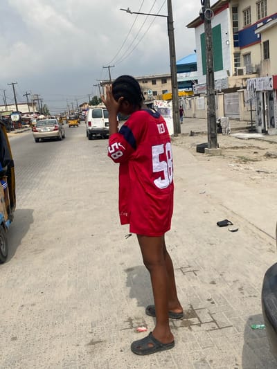 An individual stands with their back mostly to the camera on the unpaved shoulder of a two-lane road in Langbasa, Nigeria, on an overcast day. The person, wearing an oversized red jersey with white numbers "58" on the back and "RED" above it, along with dark Crocs-style footwear, has their right hand raised, partially obscuring their face. To the right, a black car bumper is visible, along with scattered debris including a black sandal. Directly behind the individual, a black pole with attached circular apparatus and wires stands on the dirt. The right side of the street features multiple commercial buildings with varying architectural styles, including a prominent light-colored building with blue trim and numerous windows, and several storefronts. Across the paved road, which shows multiple vehicles including a white van, a silver sedan, and several yellow tricycles (keke napep), utility poles with a dense network of overhead wires line the street. Further down the road, additional buildings and market stalls are visible under the gray sky.