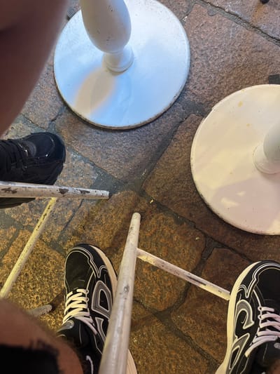 The image shows a close-up, overhead view of two pairs of legs and shoes positioned around a metal chair. One person is wearing black sneakers with white laces and white detailing. The second person is wearing a black shoe. The legs are positioned around two round, white metal bases of the chair's legs. The floor consists of uneven, stone paving. The location is Bologna, Italy, based on the pavement style and the nature of the outdoor setting.