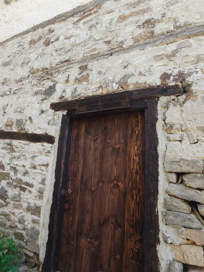 A wooden door is set in a stone wall in Kovachevitsa, Bulgaria. The door is made of dark wood with a vertical grain pattern. A dark wooden frame surrounds the door, and a wooden lintel with an inscription in Cyrillic script sits above the door. The inscription includes the word "УЧИЛИЩЕ" (School) and the years "1870-1970". The wall is constructed of irregularly shaped stones, mostly light in color, and is covered in some areas with what appears to be light-colored plaster or mortar. Green foliage is visible at the bottom left of the image.