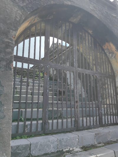 An upward-angled view captures a weathered, dark gray stone archway with a heavy, double-panel metal gate, closed and secured by a padlock, in Phú Xuân Ward, Vietnam. The gate, exhibiting significant paint chipping and rust, features vertical bars that partially obscure a long, ascending staircase beyond. The staircase, constructed of light gray stone or concrete, shows minor green vegetation growing between steps. Approximately halfway up the stairs, a small, dark-colored canine animal is visible, facing the viewer. Further in the background, atop the staircase, a multi-tiered building with traditional East Asian architectural elements and weathered facades is visible against a bright, overcast sky. The overall scene is illuminated by natural daylight.
