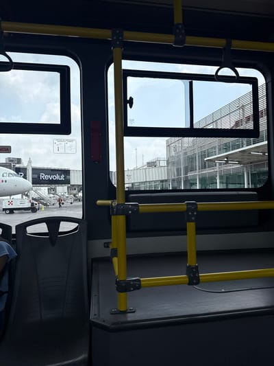 This image captures a scene from inside a public transport vehicle, likely an airport shuttle bus, looking out through its windows. The photograph is oriented vertically, showing elements both inside and outside the bus.
**Subjects & Scene:** Inside the bus, prominent yellow grab bars with black connectors stretch across the frame, indicating a typical public transport interior. Dark interior panels and a portion of a dark seat are visible in the lower right. Outside the upper window, a modern airport terminal building with a facade of glass and horizontal slatted structures can be seen under a cloudy sky. Outside the lower window, an active airport apron is visible. The front section of a white airplane, displaying what appears to be a Mexican flag, is a central feature. A jet bridge, clearly branded with the "Revolut" logo in white letters on a black background, is connected to the aircraft. Ground support equipment, including a cart with "AP-B303" printed on it, is positioned near the plane. An airport worker in an orange high-visibility vest stands on the tarmac.
**Activity:** The bus is stationary or moving slowly within an airport environment, transporting passengers or crew, or waiting on the apron. The airplane appears to be parked, possibly undergoing servicing or preparing for boarding/deplaning.
**Notable Details:** The scene takes place during the daytime, under a partly cloudy sky. Location cues such as the airplane, jet bridge, ground crew, and terminal building firmly place the setting at an airport, specifically in Apodaca, Mexico, as per the context.
**Visible Text:** "Revolut" is clearly visible on the jet bridge. On one of the bus windows, "VENTANA DE EMERGENCIA" (Emergency Window) and a corresponding instruction diagram are present. Text "AP-B303" can be seen on the ground support equipment.
