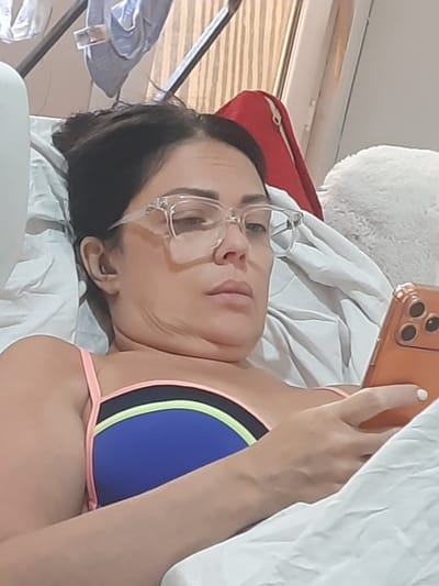 A woman with dark hair and glasses is lying down, presumably on a bed, while holding a smartphone. She is wearing a blue and neon green bikini top with pink straps. The background includes a white pillow, a red object, and a window with blinds. The exact location in Al Hadaba, Egypt cannot be determined from this image.